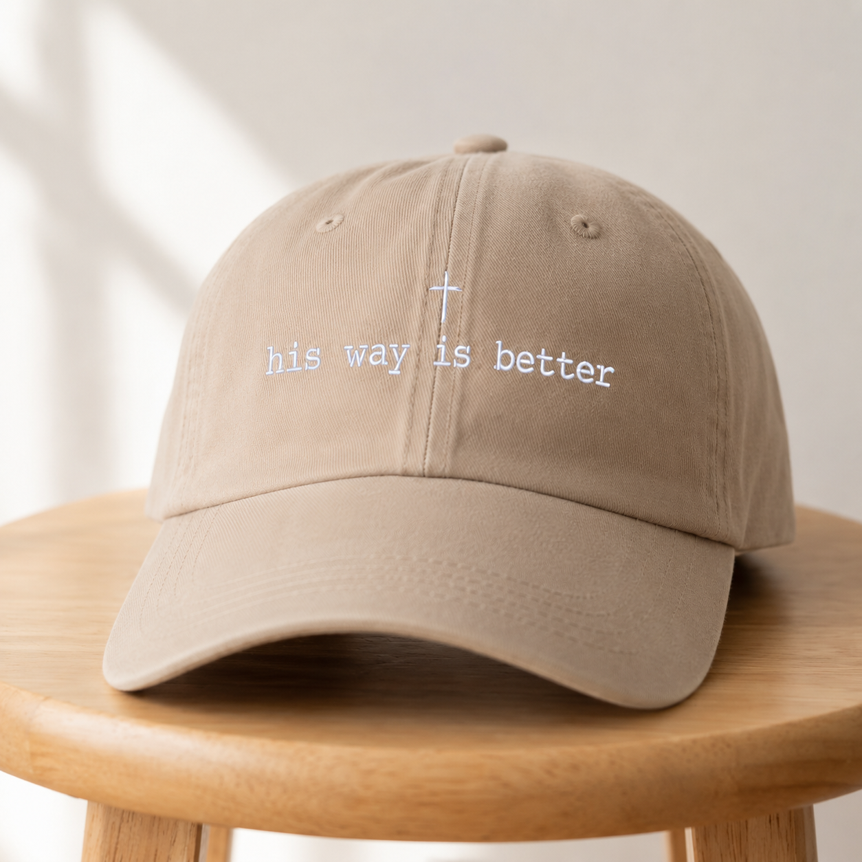 A beige baseball cap with white embroidered text reading 'his way is better' and a small cross, displayed on a round wooden stool against a softly lit neutral background.