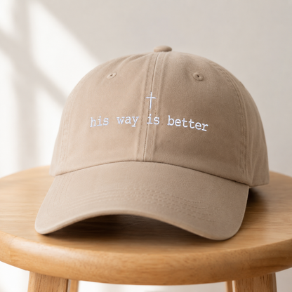 A beige baseball cap with white embroidered text reading 'his way is better' and a small cross, displayed on a round wooden stool against a softly lit neutral background.