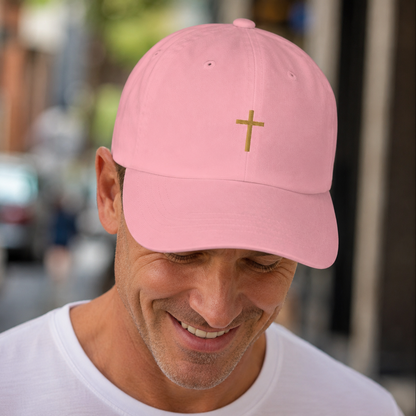 A man wearing a light pink baseball cap with a small gold cross embroidered on the front, photographed outdoors with a blurred street background. Best Christian hat and Christian gift