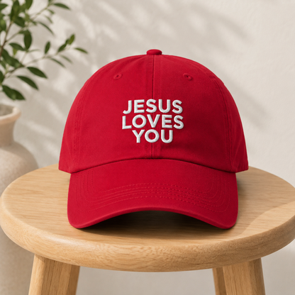 A red baseball cap with white embroidered text reading 'JESUS LOVES YOU' sits on a light wood stool. The background is a warm beige wall with soft leaf shadows, and a blurred plant and vase are visible on the left.