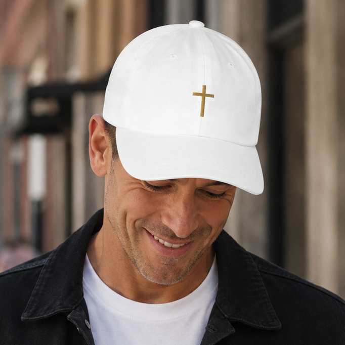 A man wearing a white baseball cap with a small gold cross on the front, a white t-shirt, and a black denim jacket, walking down a city street. Best Christian cap and faith gift ever