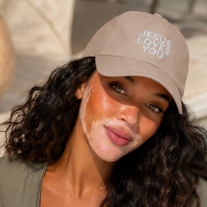 A woman with dark curly hair wearing a beige baseball cap that reads 'JESUS LOVES YOU' and an olive green ribbed crop top, posing in front of a swimming pool.